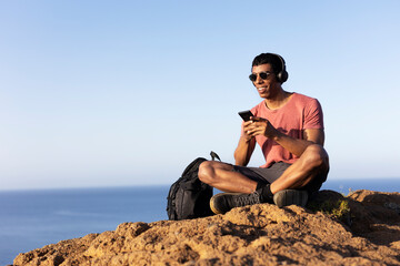 Young man sitting on the stone listening the music. Tourist man on the top of the mountain