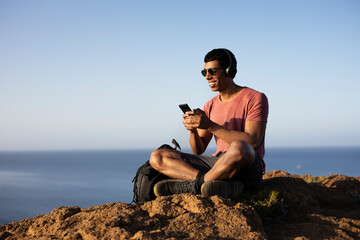 Young man sitting on the stone listening the music. Tourist man on the top of the mountain