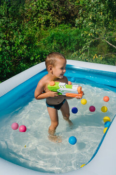 Kids Play With Water Gun Toy In Garden. Little Boy Playing With Water Pistol Hose In Sunny Backyard. A Happy Child Is Splashing And Swimming In An Inflatable Pool. Kid Having Fun.