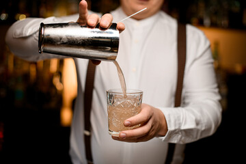 close-up of bartender holds crystal glass into which pours drink from shaker cup
