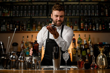 various shakers and bottles stand on the bar counter and male bartender gently pours alcoholic drink into cup