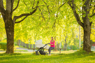 Obraz na płótnie Canvas Young adult woman sitting on wooden bench between trees at town park in spring day. White baby stroller beside mother. Relaxing after long walk. Side view. Peaceful atmosphere.