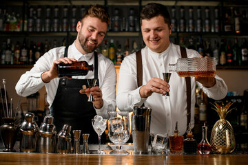 Two cheerful bartenders pouring a drink from a steel jigger bottle.