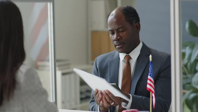 African American visa officer taking documents from female applicant and speaking with her during interview in US embassy