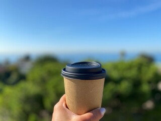 Woman's hand holding a recycled paper coffee cup at the coast on a sunny day showing trees, sea and sun