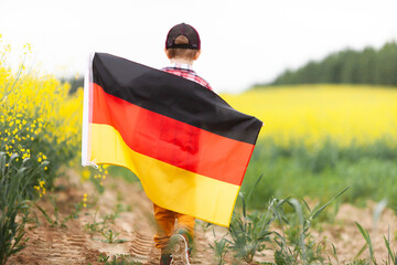 Little boy walking holding flag of Germany in the rapeseed field. Back view. Day of German Unity....