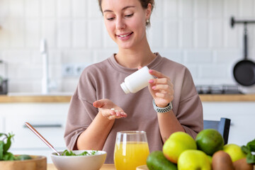 Vitamins, nutritional supplements, healthy lifestyle concept. Young woman eating breakfast and taking vitamin sitting at home in the kitchen, smiling friendly