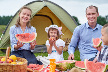 Happy family eating watermelon at picnic in meadow near the tent. Mother, father and child Enjoying Camping Holiday In Countryside