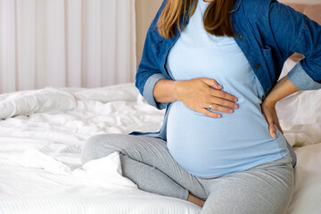 Pregnant woman touch her tummy and keep one hand on her back sitting on bed in her bedroom. Back pain, backache, labor pains, healthcare, pregnancy health problems.
