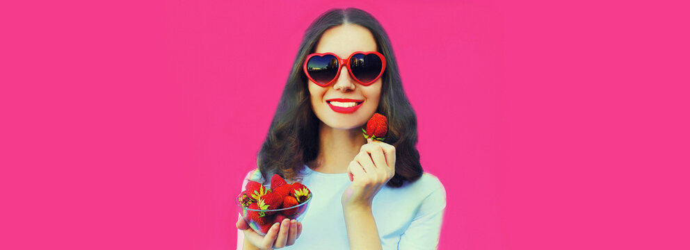 Portrait close up of happy smiling woman with handful of fresh strawberries wearing red heart shaped sunglasses on pink background