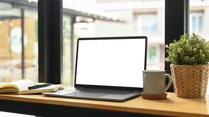 Fototapeta premium Computer laptop with blank display, potted plat, coffee cup and notebook on wooden table