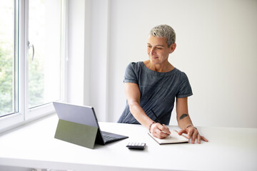 middle-aged businesswoman standing by white high table and working on tablet and wearing grey-blue dress and with short light hair