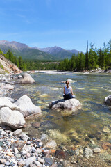 A girl in a yoga pose sits on a stone on the river bank and meditates looking at the mountains on a sunny summer day. Rest at nature. Summer travel. Beautiful landscape. Natural background