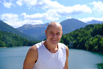 Naklejka na ściany i meble mature man of 60 years old stands, smiles against backdrop of lake Achensee in Austria, green mountains rises above calm expanse of water, concept of vacation by reservoir, resort place tyrol
