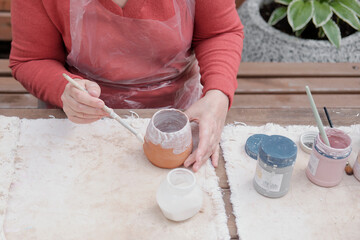 Hands of older woman painting handmade clay vase with white paint with brush.