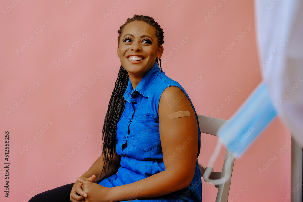 Wall mural smiling black young adult woman with bandage plaster on arm sitting on chair after vaccination looking at doctor studio shot