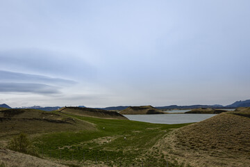 A lake behind grass covered hills with Icelandic sheep grazing on them. Volcano and mountain range on the horizon 