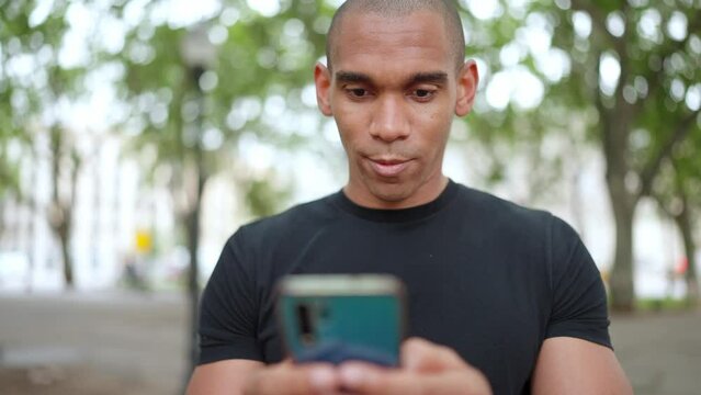 Positive bald African wearing black t-shirt typing on phone while sitting on the bench outdoors 