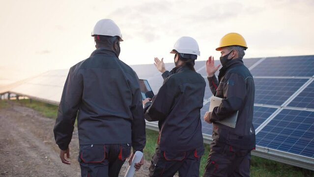 Three engineers in protective face masks, hard hats and uniforms walk among rows of solar panels and communicate. View from back.