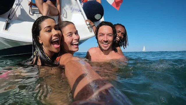Happy multiracial friends streaming live video swimming in the sea with sail boat in background 