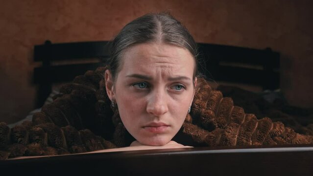 Young girl is sad lying on a bed under a blanket close-up. he face of a beautiful girl looks sadly and dejectedly at one point, as if she is about to cry. Disorder.