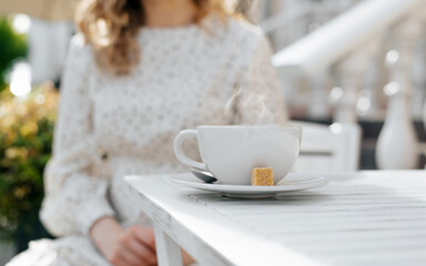Breakfast coffee in street cafe. Close-up of an unrecognizable woman sitting at table with cup of drink outdoors on sunny morning. Selective focus on white cup