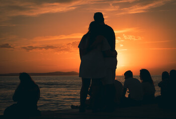 Zakochana para przytula się i patrzy na zachód słońca. Ludzie siedzący na promenadzie patrzący na zachód słońca. Zdjęcie pod światło,  zarys  sylwetek. - obrazy, fototapety, plakaty