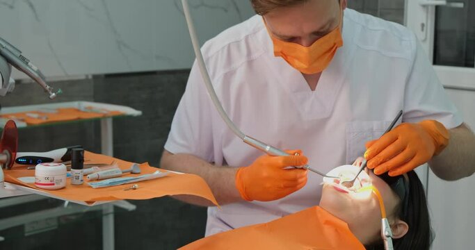 dental clinic, proficient stomatologist fellow in orange gloves and mask professional cleaning oral cavity for patient who with dilator in mouth lies in medical chair in stomatology hospital