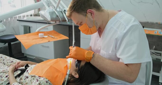 dental health, proficient stomatologist fellow in orange gloves and mask makes polishing teeth procedure for patient who with dilator in mouth lies in medical chair in hospital