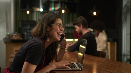 Happy girl staff talking on phone smiling and laughing browsing online orders sitting at coffee shop table