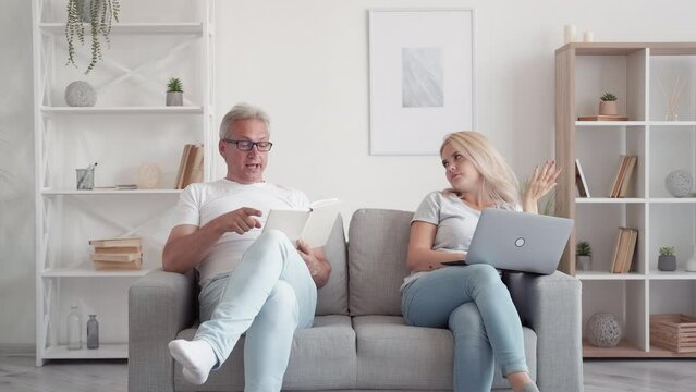 Family Mentoring. Old-fashioned Education. Modern Technology. Conservative Middle-aged Dad With Book Speaking To Annoyed Daughter Studying Using Laptop On Couch At Home Interior.