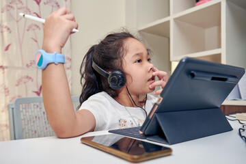 Little preschool girl with headphones using digital tablet computer for online education at home office.