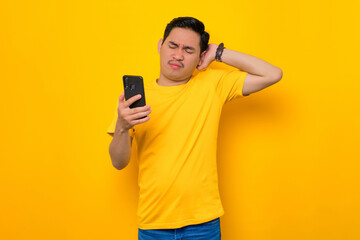 Confused young Asian man in casual t-shirt using mobile phone while scratching head, reacting to online news isolated on yellow background. People lifestyle concept