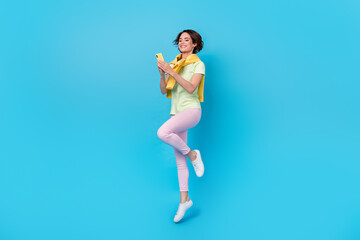 Fototapeta na wymiar Full size photo of cute millennial brunette lady jump hold telephone wear t-shirt pants footwear isolated on blue background