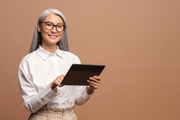 Modern elderly senior woman in formal wear using digital tablet isolated on beige. Portrait of...