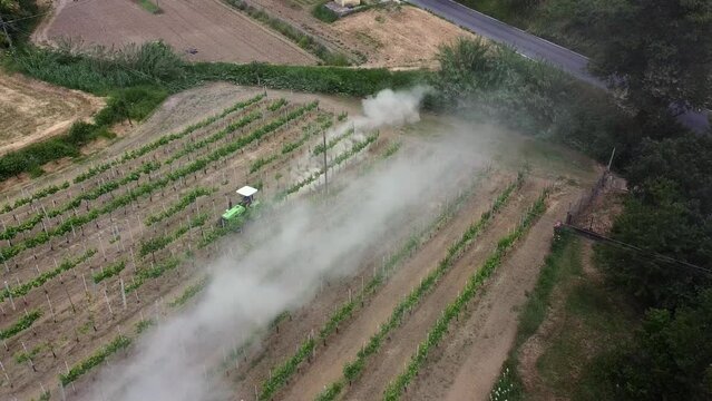 Aerial shot of a compact tractor hidden in the dust as it ploughs the ground between the existing vines