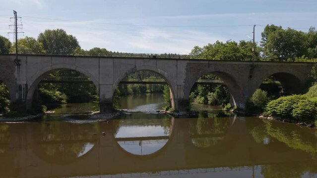 Young man standing on a footpath underneath a train bridge that reflects perfectly in the flat brown river water. Low aerial fly through shot