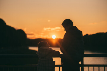 
silhouettes of a young man in a dark jacket with a little girl - a sister in a beige coat, talking and standing at sunset on a city bridge over a river, medium close-up