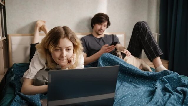 The couple is in bed in the morning. A young woman points at something to a guy on a laptop. A young man pets a dog. The woman smiles. Slow motion 4k footage