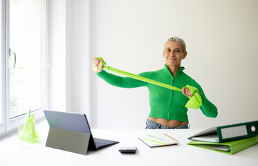 middle aged businesswoman standing at white high table in office doing stretching exercises with green stretch band or gymnastic band