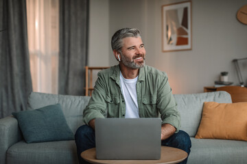 Smiling elderly european man with beard in wireless headphones typing on laptop and looking at empty space