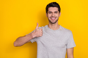 Photo of funny brunet young guy show thumb up wear grey t-shirt isolated on yellow color background