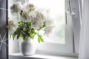 A beautiful white bouquet of peonies in the sun on the windowsill. Flowers and buds in a vase. Light, white background.