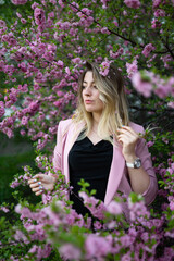 A very beautiful blonde in a pink jacket and a black blouse sensually poses standing among the flowers of cherry blossoms. Goddess of nature or earth