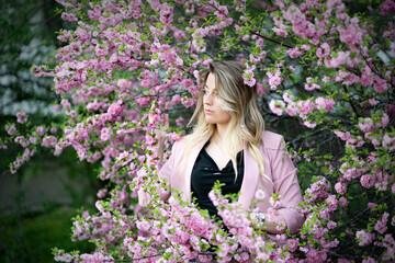 A very beautiful blonde in a pink jacket and a black blouse sensually poses standing among the flowers of cherry blossoms. Goddess of nature or earth