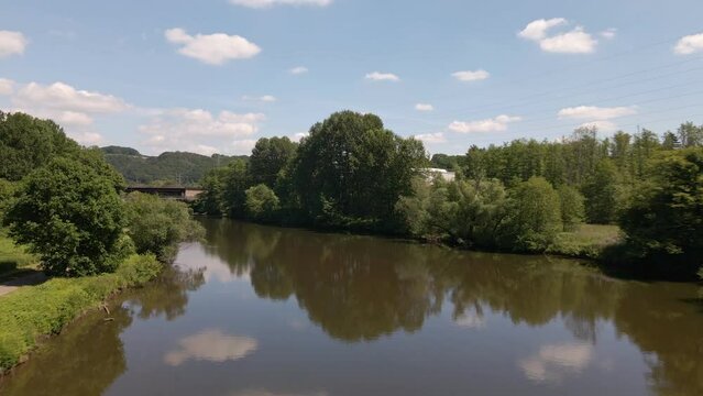 Blue sky and lush trees reflecting in the brown water of a calm river in west Germany. Wide angle aerial flyover