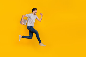 Fototapeta na wymiar Full size profile photo of funny brunet young guy run with laptop wear t-shirt jeans shoes isolated on yellow background