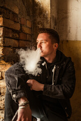 Portrait of a brutal smoking macho man in a black denim jacket with a stylish hairstyle, piercing in the eyebrow and lip and an earring in the ear. A rock musician or punk in an abandoned building