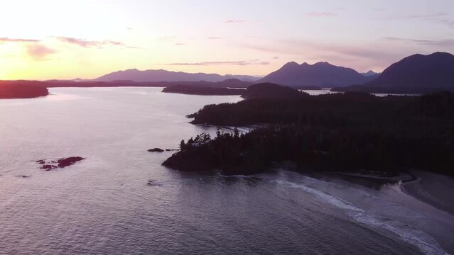 A panning drone video over the ocean next to a forested coast during sunset with mountains visible in the background.