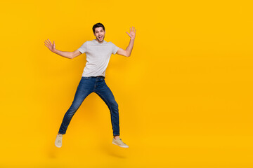 Fototapeta na wymiar Full size photo of cool brunet young guy run jump wear t-shirt jeans footwear isolated on yellow background